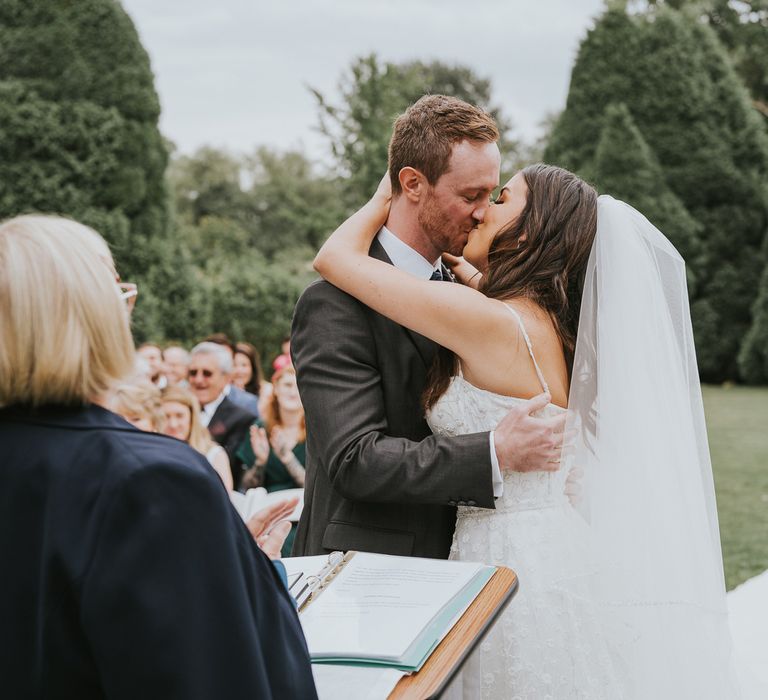 Bride in white cami strap Pronovias wedding dress and veil kisses groom min grey three piece suit during outdoor wedding ceremony at Notley Abbey