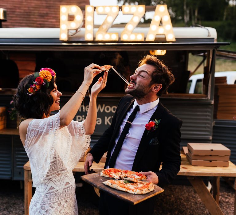 Bride in strapless Grace Loves Lace wedding dress feeds groom in black suit a slice of Pizza by pizza van at rustic barn wedding reception