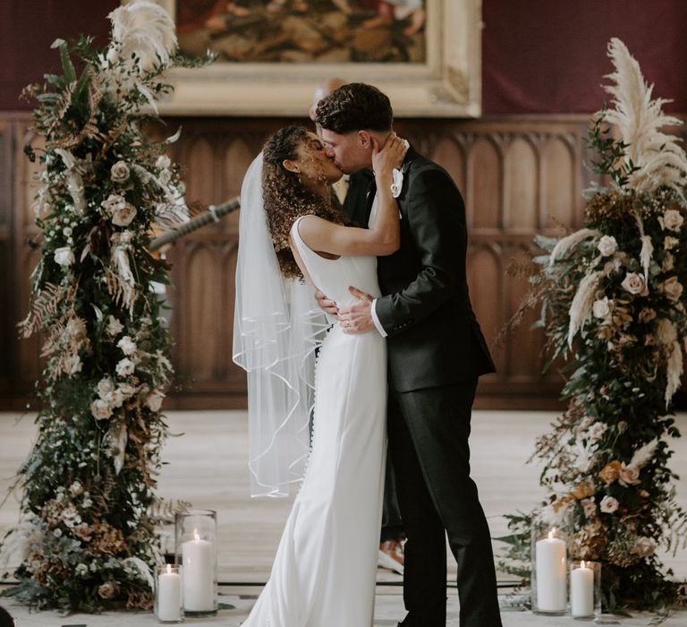 Bride & groom kiss as they stand in front of floral archway 