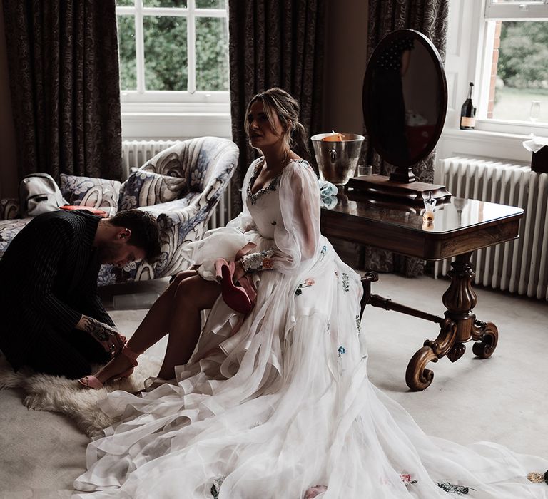 Bride with ponytail wearing long sleeve Victoria Sanders wedding dress with train and floral embroidery sits on stool whilst having her shoe tied