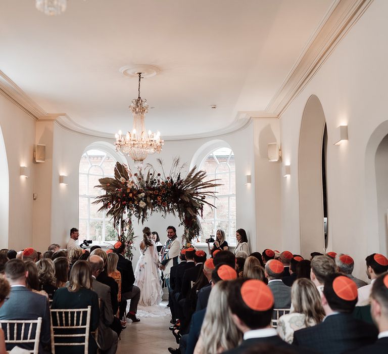 Bride in long mesh sleeve wedding dress with floral embroidery stands with groom in kippah and green suit under arch with dried flower cloud for interfaith wedding ceremony at Iscoyd Park