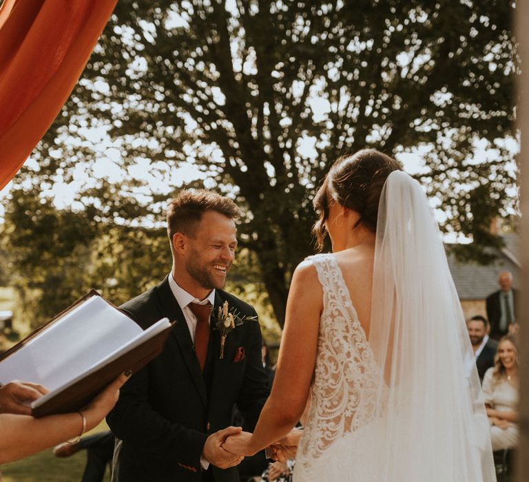 Bride in lace low back wedding dress and veil stands holding hands with groom in dark suit and burgundy tie during outdoor wedding ceremony at Hope Farm in Dorset