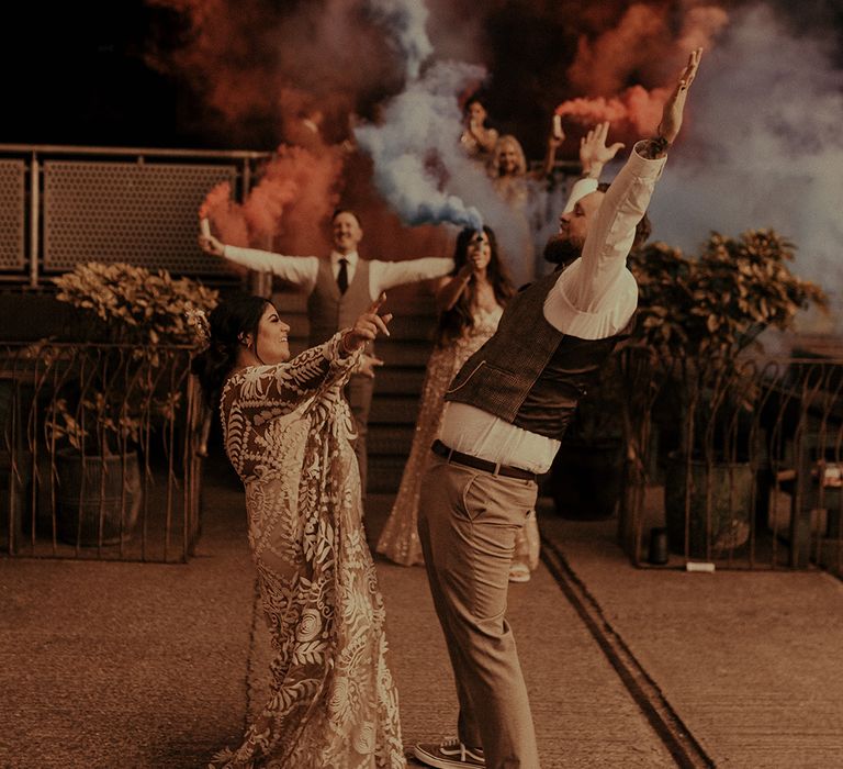 Wedding party holding smoke flares whilst the bride and groom dance in a waistcoat and trainers and boho wedding dress 