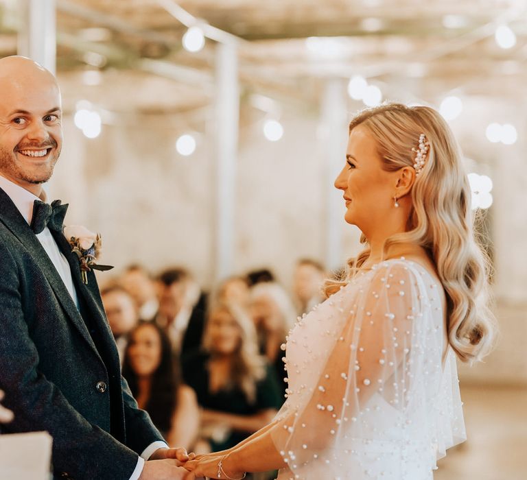 Bride in pearl wedding dress with pearl drop earrings and pearl hair slider stands with groom in navy tweed suit with bow tie and buttonhole during wedding ceremony