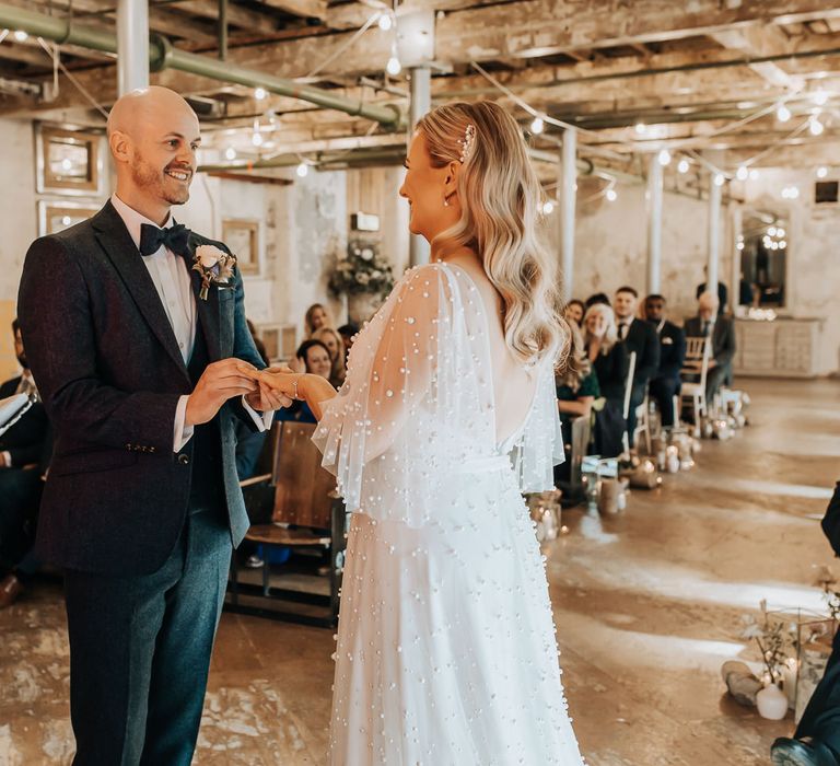 Groom in navy suit and bow tie puts ring on finger of bride in low back pearl wedding dress with sleeves and pearl hair clip during wedding ceremony