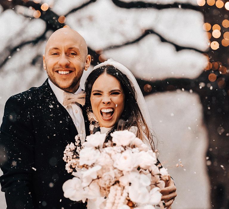 Groom in a black suit and bride in a long sleeve wedding dress holding an all-white wedding bouquet at snowy winter wedding with fairy lights 