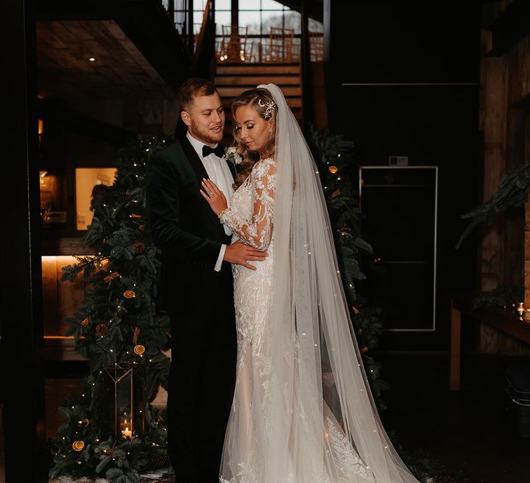 Bride in lace wedding gown and groom in black tie stand in front of dried orange decorated stairway on wedding day