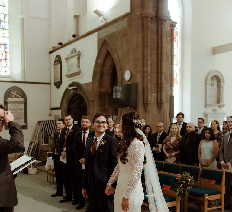 Bride in lace gown and groom in blue suit smile at each other at the altar of their church wedding ceremony