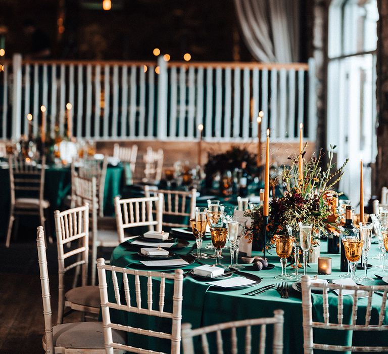 Cosy wedding venue with exposed lightbulbs and tables with green tablecloth, yellow candles and glasses and red and orange flower centrepiece 