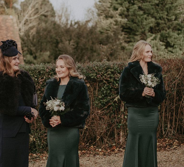 Bridesmaids in dark and bottle green bridesmaids dresses with mixed floral bouquets, black cropped fur jackets and black hats