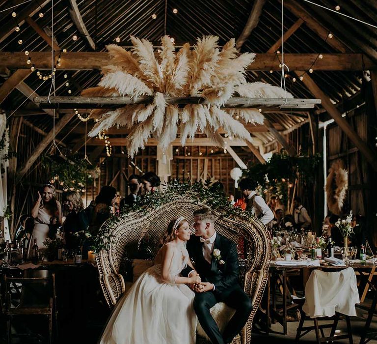 Rustic barn wedding with bride and groom sitting on a peacock chair under a giant disco ball and pampas grass installation 