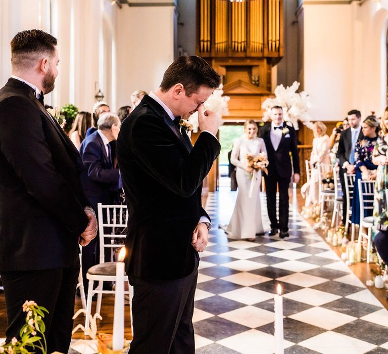 Groom in black tie cries as the bride walks down the aisle with her father leading her 