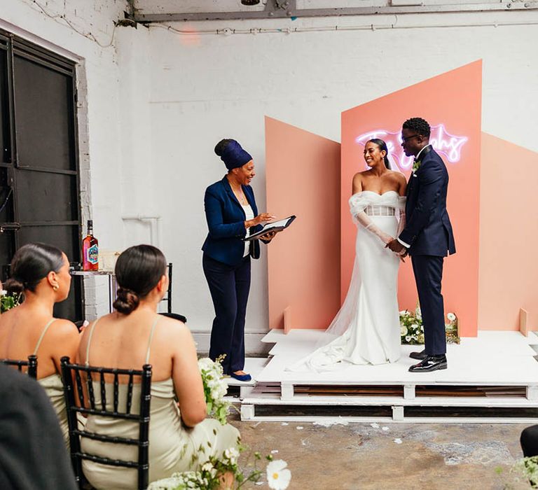 Bride and from stand on raised white platform with pink background and white neon light at industrial venue