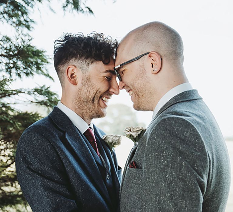 Grooms laugh and rest their foreheads together on their wedding day wearing dark blue and grey suits with paper rose buttonhole