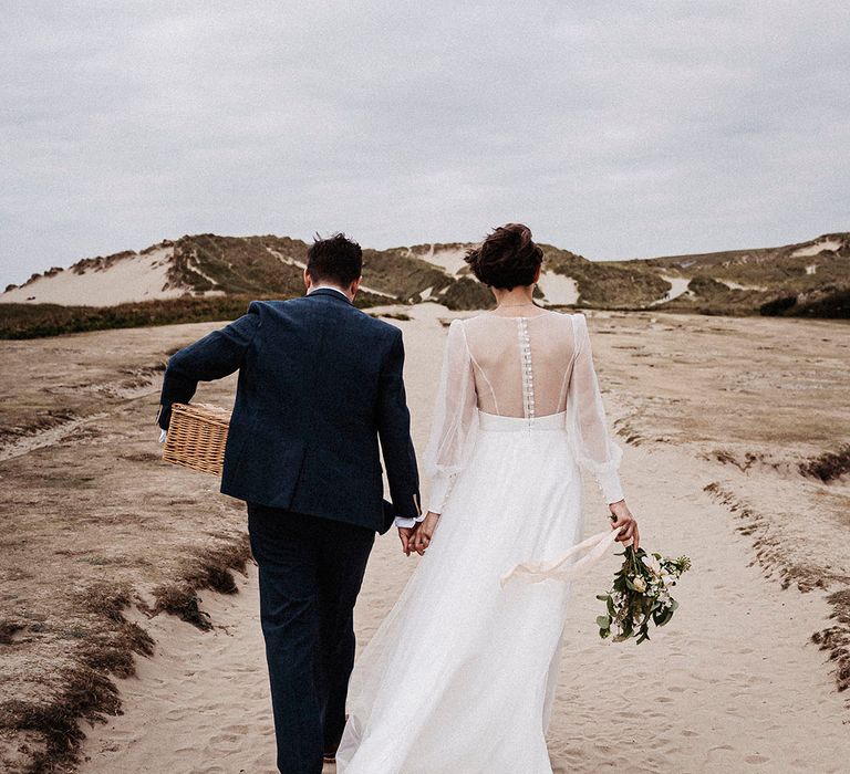 Bride and groom walk along the beach at Cornwall during their wedding day with the groom carrying a wicker picnic basket and the bride holding a wedding bouquet 