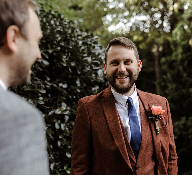 Groom in three piece brown suit with pink rose buttonhole and blue tie speaks to wedding guest