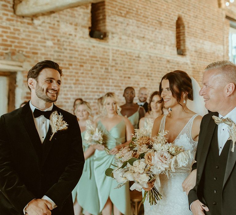 Father of the bride walks the bride to the end of the aisle where the groom waits in black tie and as they all smile at each other