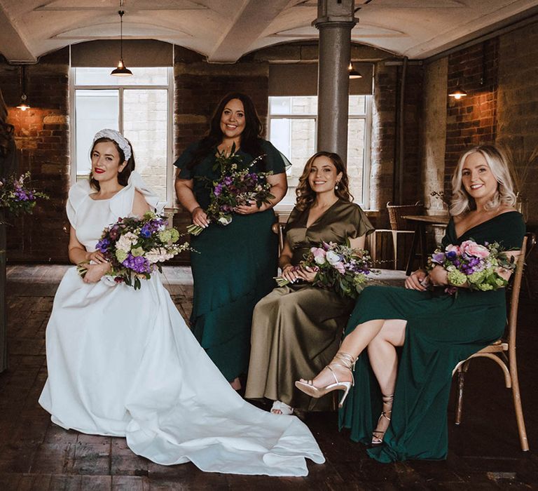 Bride in Jesus Peiro wedding dress with bridesmaids in sage, emerald, and olive dresses in various styles as they all hold purple wedding bouquets