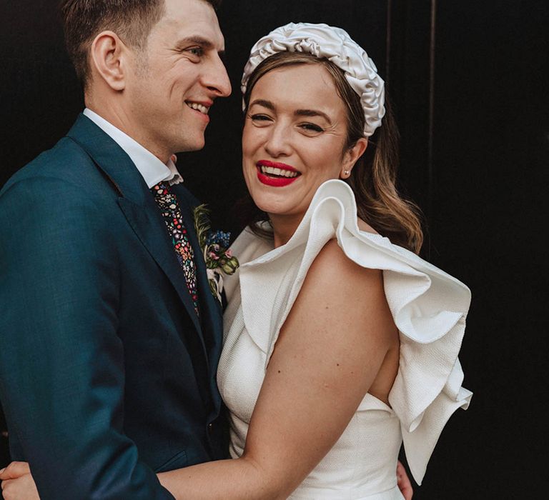 Smiling bride with red lipstick, white ruffle headband and wedding dress with statement sleeves and smiling groom in navy blue suit and floral tie