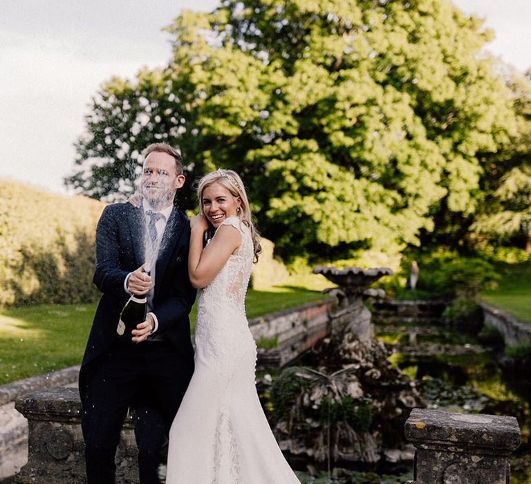 Groom in morning suit pops a bottle of champagne with bride leaning against him in lace wedding dress and pearl headpiece 