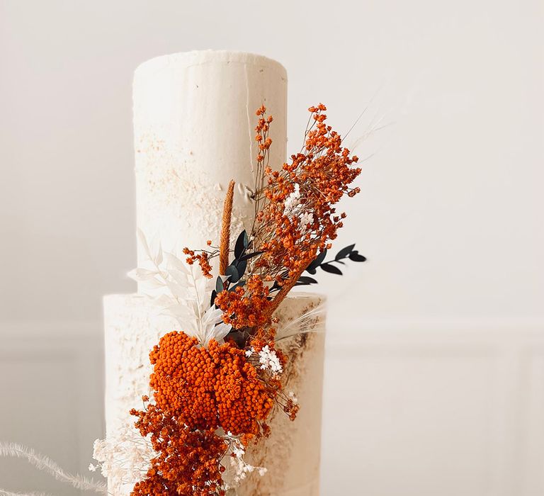 Tall white three tier wedding cake with textured icing decorated with orange, white and black dried flowers and grasses 