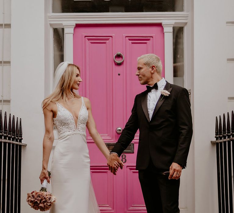 Bride and groom at each other in the eyes as they stand holding hands in front of pink painted door 