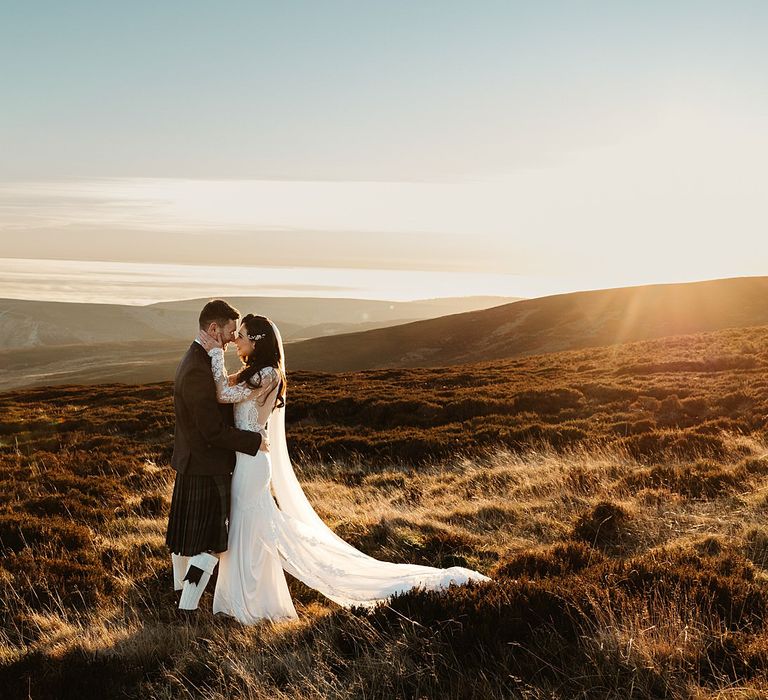 Bride & groom stand on hillside in front of beautiful Scottish highlands whilst the sun sets 