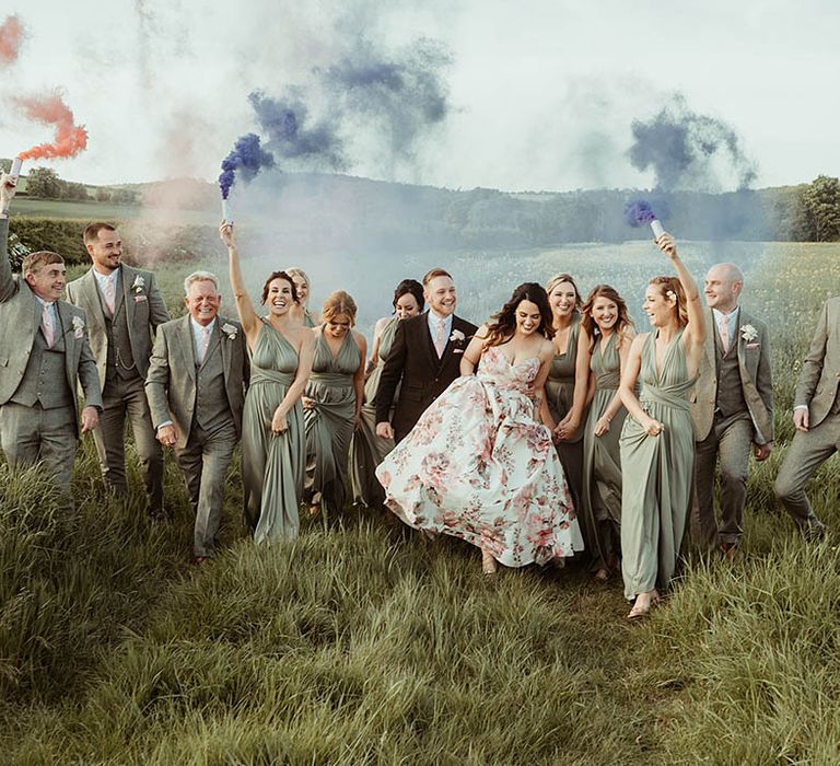 Wedding party shot with bridesmaids and groomsmen in green outfits waving colourful smoke bombs with the bride and groom 