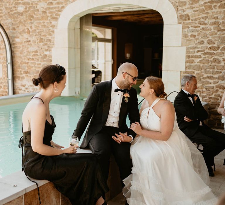 Bride wearing London Halfpenny wedding dress sits beside her groom in black tie and wedding guest in black silk dress beside pool