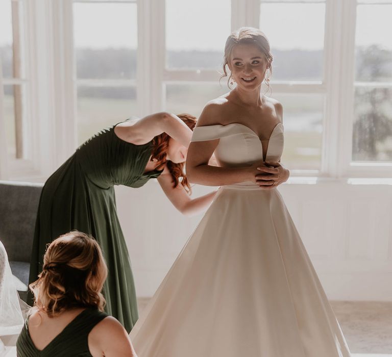 Bridesmaids in deep green dresses help the bride into her wedding dress 