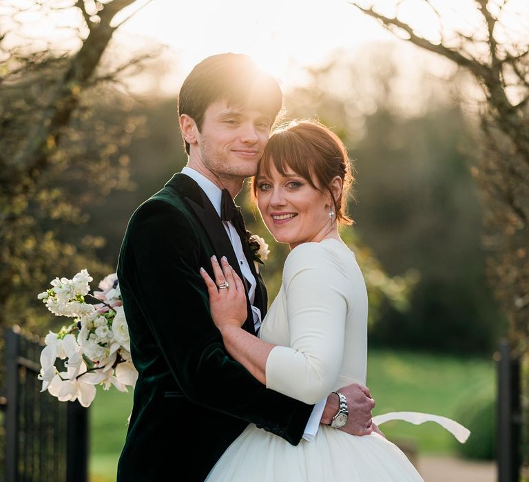 Bride wears tulle skirt princess wedding dress whilst stood beside her groom in velvet green jacket with black tie 