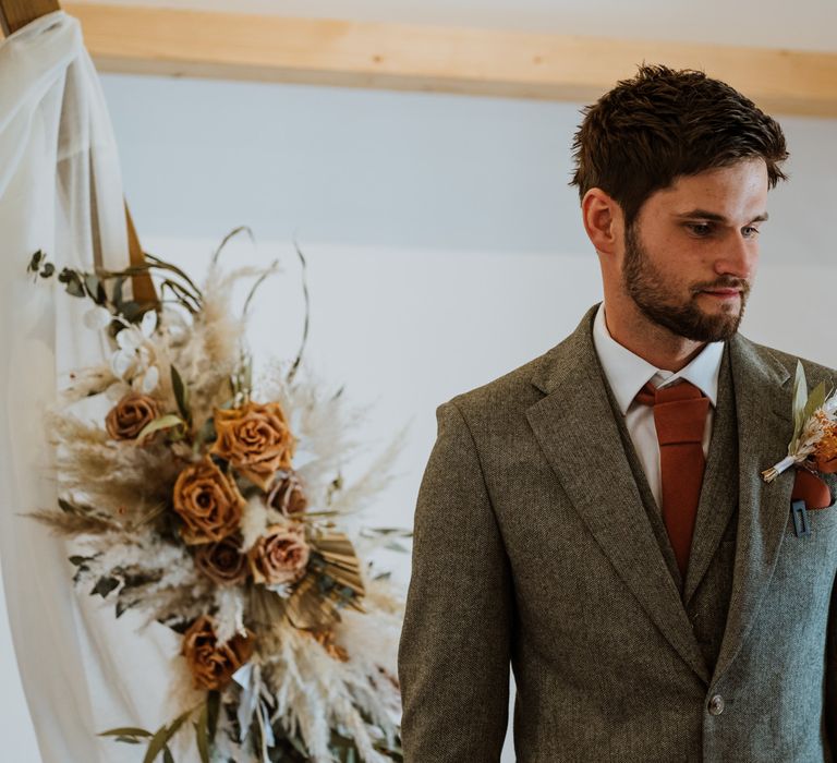 Groom in grey suit with orange tie, orange pocket square and orange and dried flower boutonniere standing by large wooden arch with a dried flower arrangement on top 