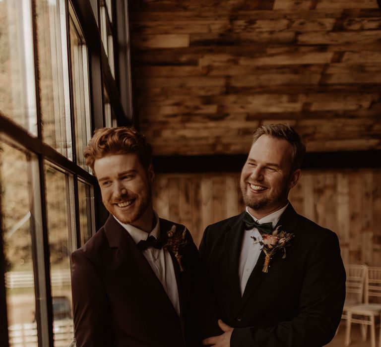 Grooms stand with linked hands looking out the window at Hidden River Cabins wedding venue 