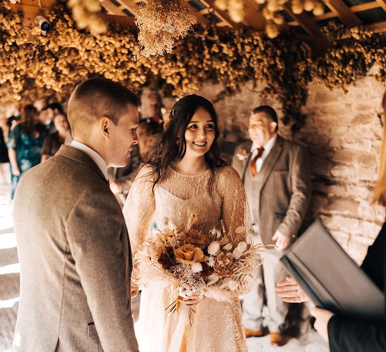 Bride holds Autumnal styled dried floral bouquet whilst wearing sheer gold wedding dress 