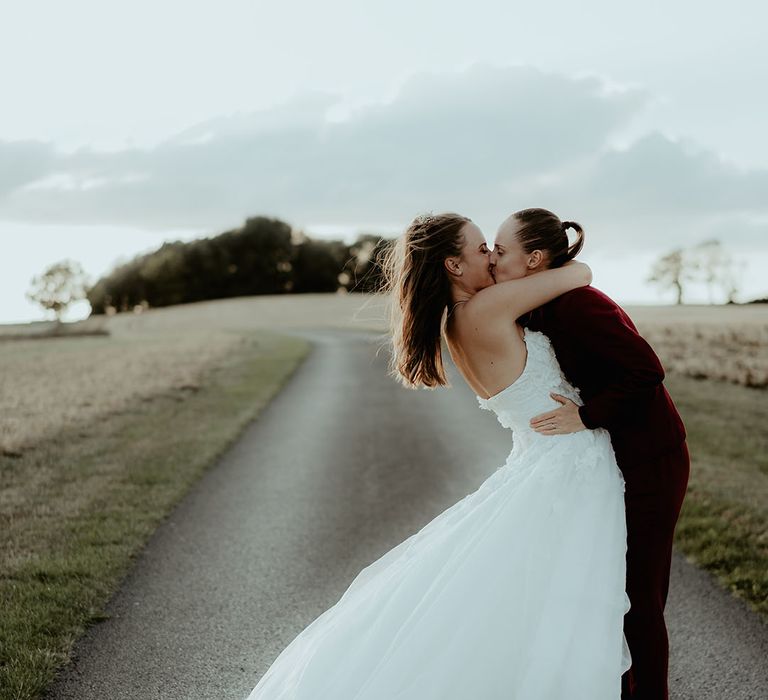 Bride in a burgundy suit and black shirt kisses the bride in a floral wedding dress 