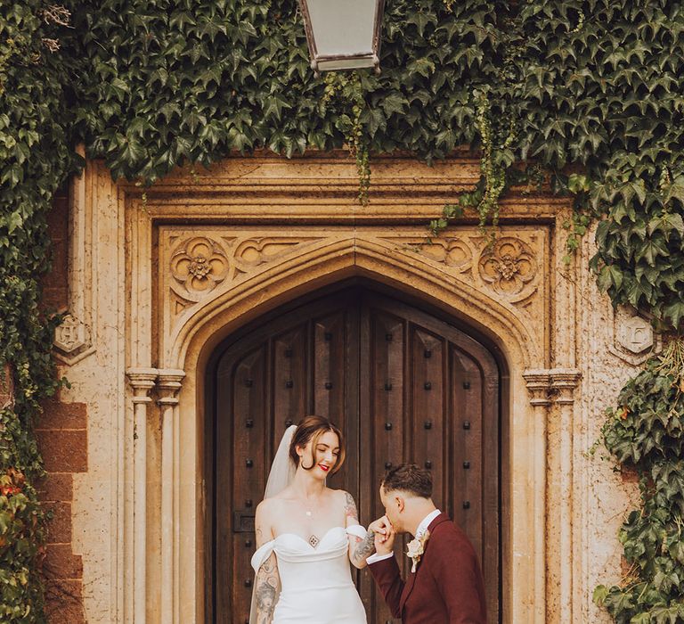 Groom in maroon suit kisses his brides hand outside St Audries Park on their wedding day