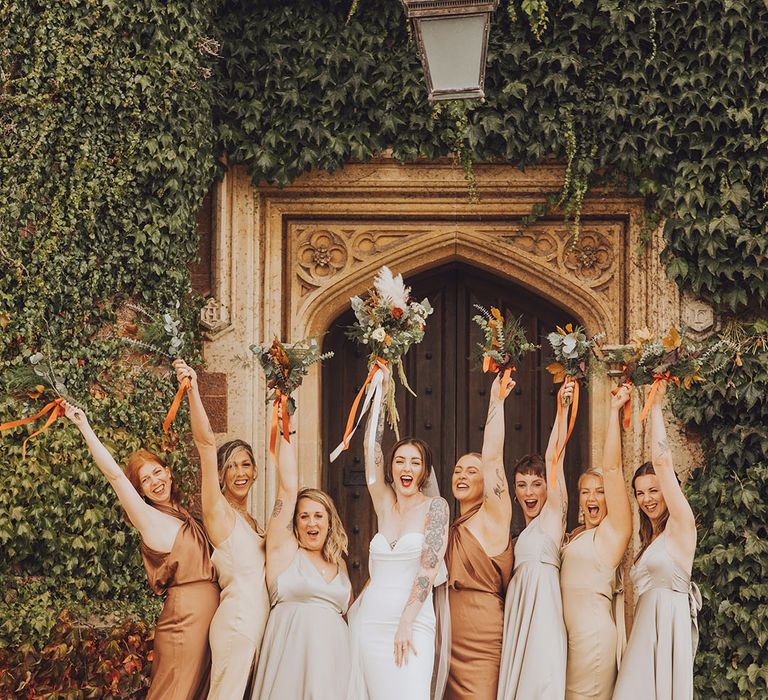 Bride lifts floral bouquet in the air with her bridesmaids in Autumnal bridesmaid dresses in mismatched styles