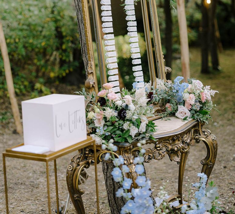 Seating plan on gold antique mirror surrounded by carnations, thistles, roses and wildflowers nest to a white love letters box