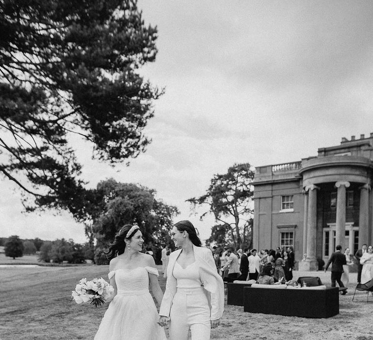 Bride in an off the shoulder wedding dress walking hand in hand with the bride in a white bridal suit 