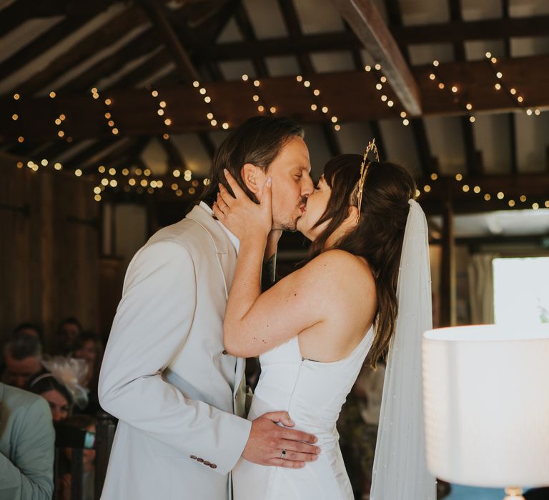 Bride in silk-look v-neck sleeveless ASOS wedding dress and gold halo headband with sparkly stars and veil kissing groom in double breasted ASOS suit at the alter