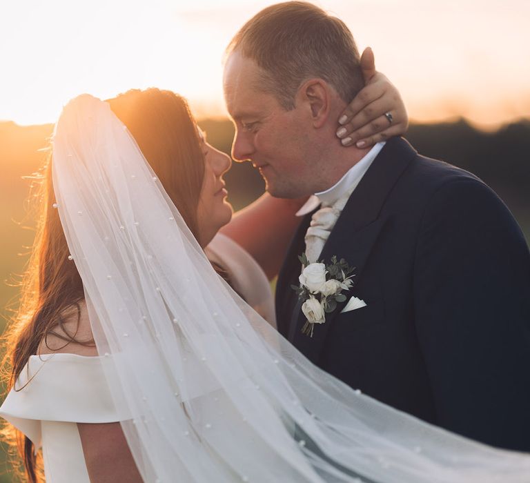 Bride wearing a pearl wedding veil resting her hand on the back of the groom's head during golden hour