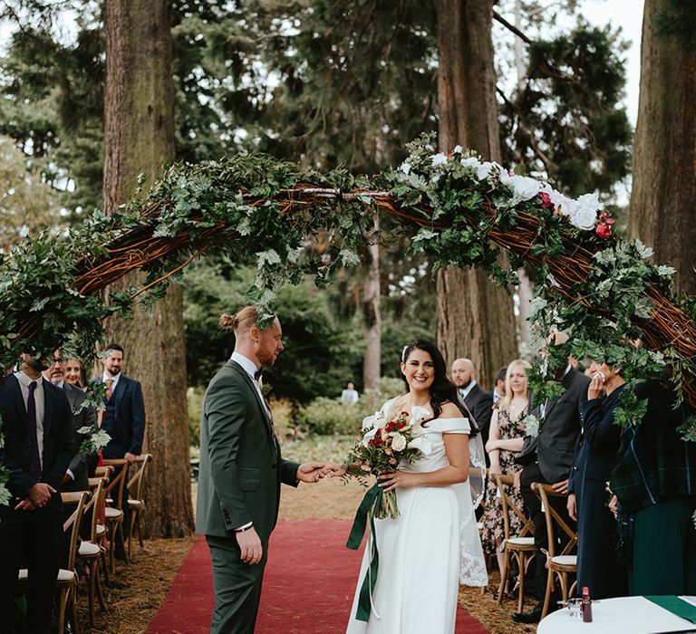 Groom in green suit & bride stand beneath green foliage archway during outdoor wedding ceremony in Edinburgh