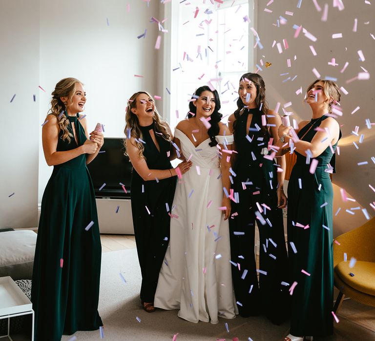 Bridesmaids in high-neck green bridesmaid jumpsuits pop confetti canons beside bride 