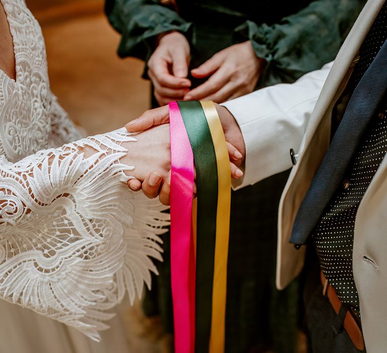 Bride in long sleeve boho lace wedding dress and groom in white blazer doing handfasting ceremony with pink, forest green and mustard coloured ribbons 