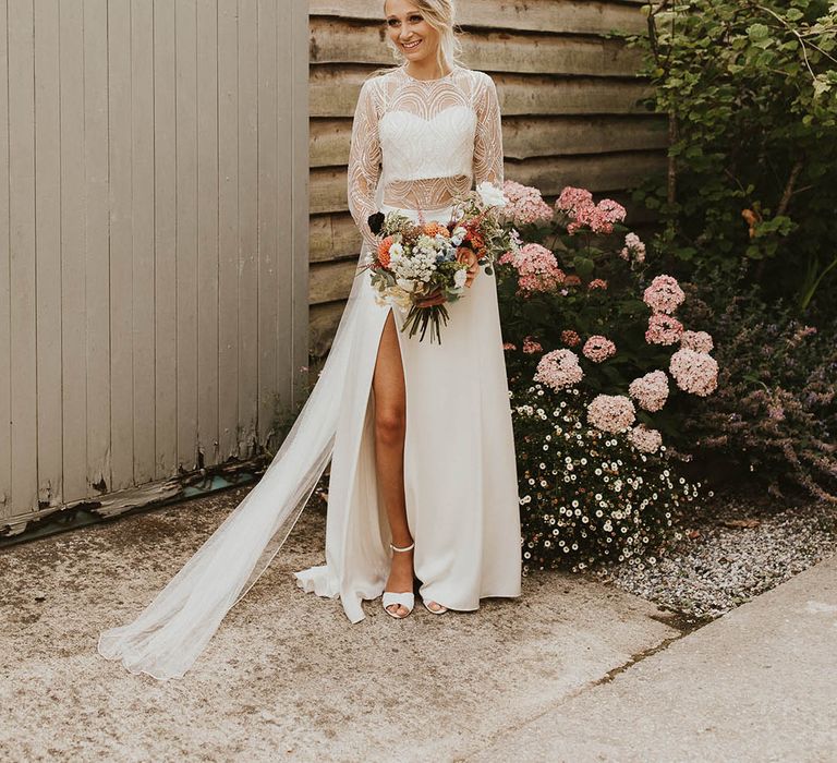 Bride in Catherine Deane wedding dress separates with lace top and side slit skirt holding an orange dahlia wedding bouquet 