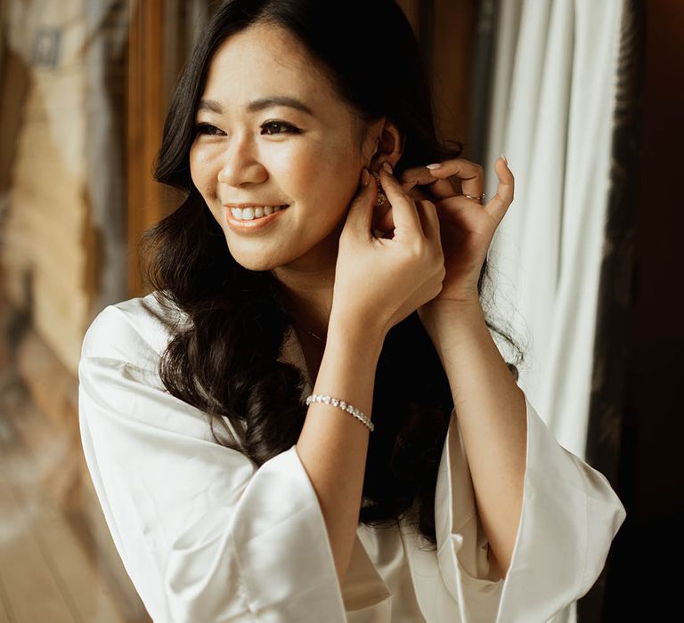 Bride wearing a diamond tennis bracelet in a white satin robe puts in her earrings for her wedding day 