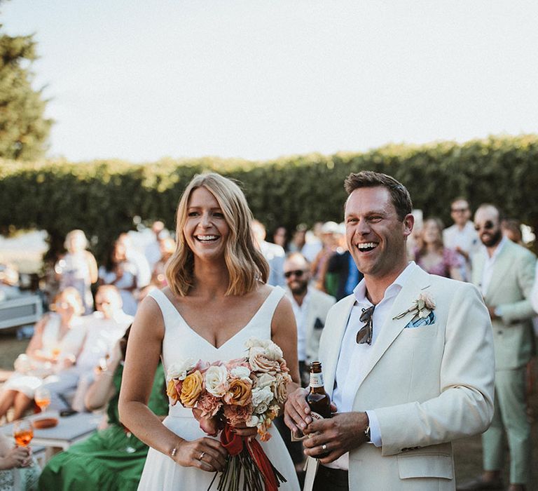 Bride holds pastel rose wedding bouquet and stands beside her groom in cream coloured suit 