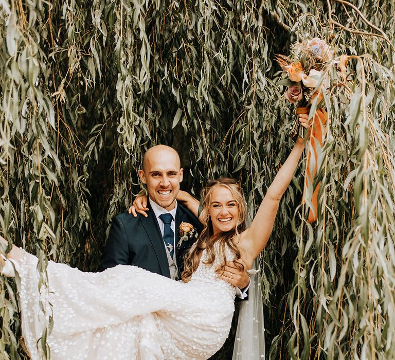 The bride in a navy suit lifts the bride up in front of willow tree at Duddon Mill Farm Cheshire wedding venue