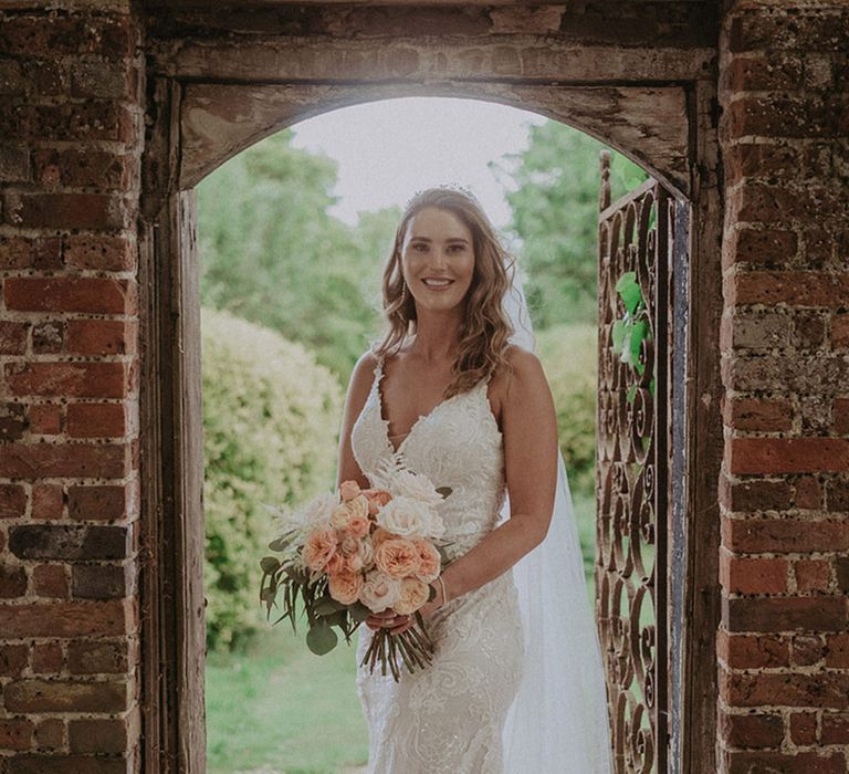 The bride wears a fitted lace and embellished bridal gown with tiara and cathedral length veil holding a peach and white rose bridal bouquet