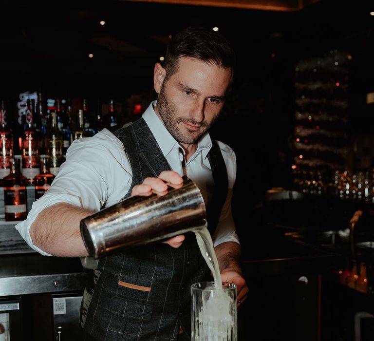 Groom in dark grey checkered waistcoat, trousers, and white shirt with no tie mixes up some cocktails for the wedding 